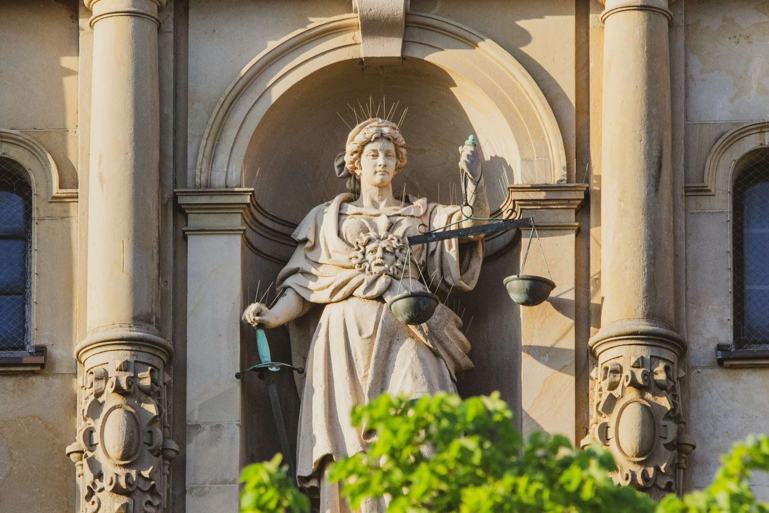 A statue of justice in Hamburg. A figure sits in an alcove, holding a set of discoloured copper and brass scales in her left hand, and her right hand resting on a sword. She looks towards the scales. She is flanked by two columns and the top branches of a